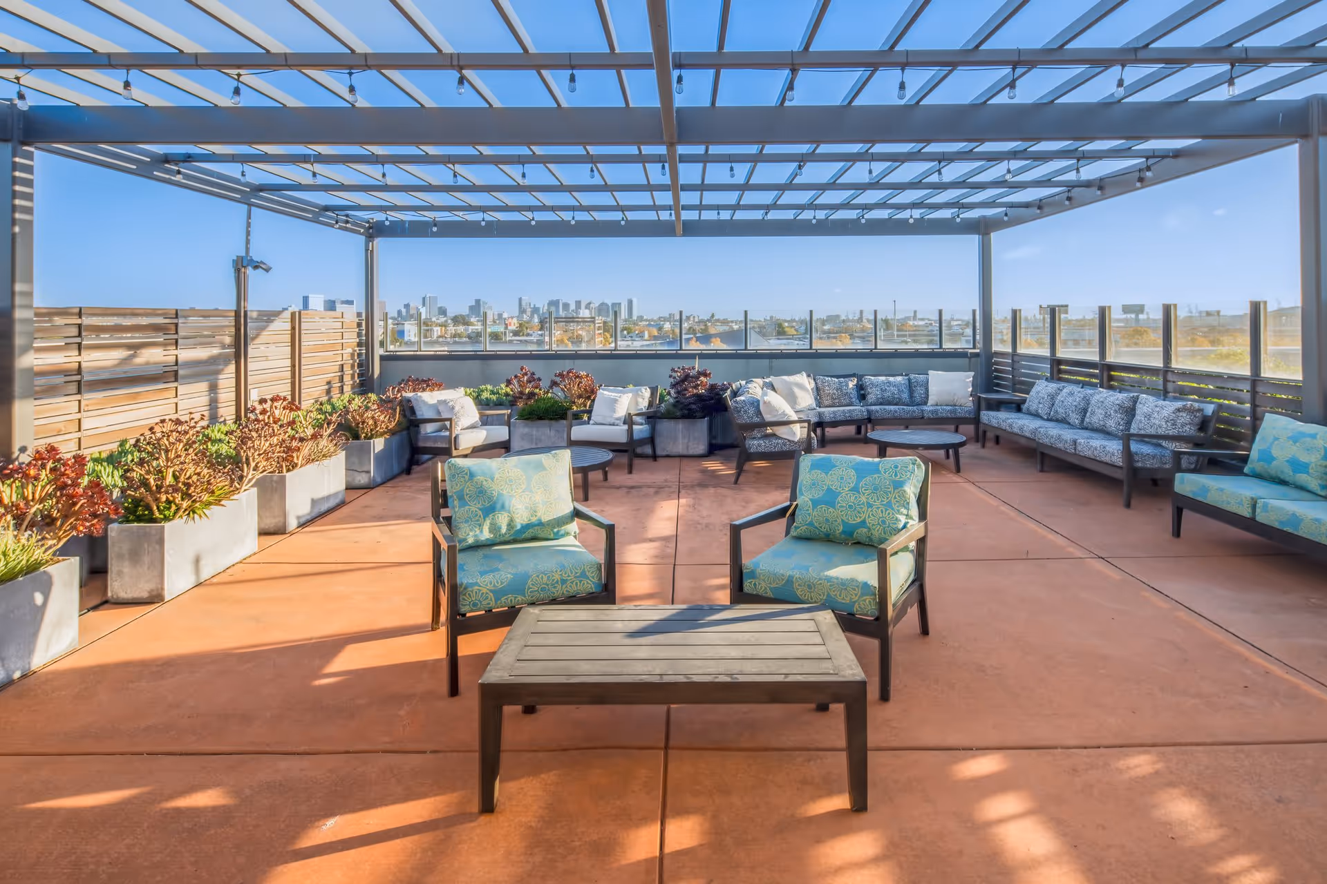 Outdoor patio area with a pergola overhead, featuring multiple cushioned chairs and sofas arranged around coffee tables. There are planter boxes with greenery along the sides and a city skyline visible in the background under a clear blue sky.