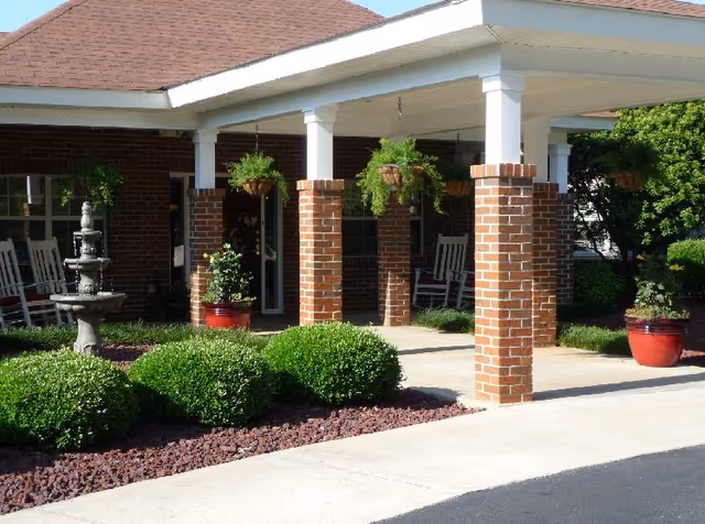 Covered entrance with brick columns, hanging plants, benches, and a fountain at Autumn Cove Assisted Living.