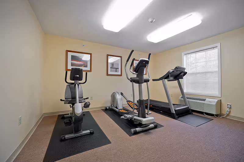 Small fitness room with a recumbent bike, elliptical trainer, and treadmill under fluorescent lights.