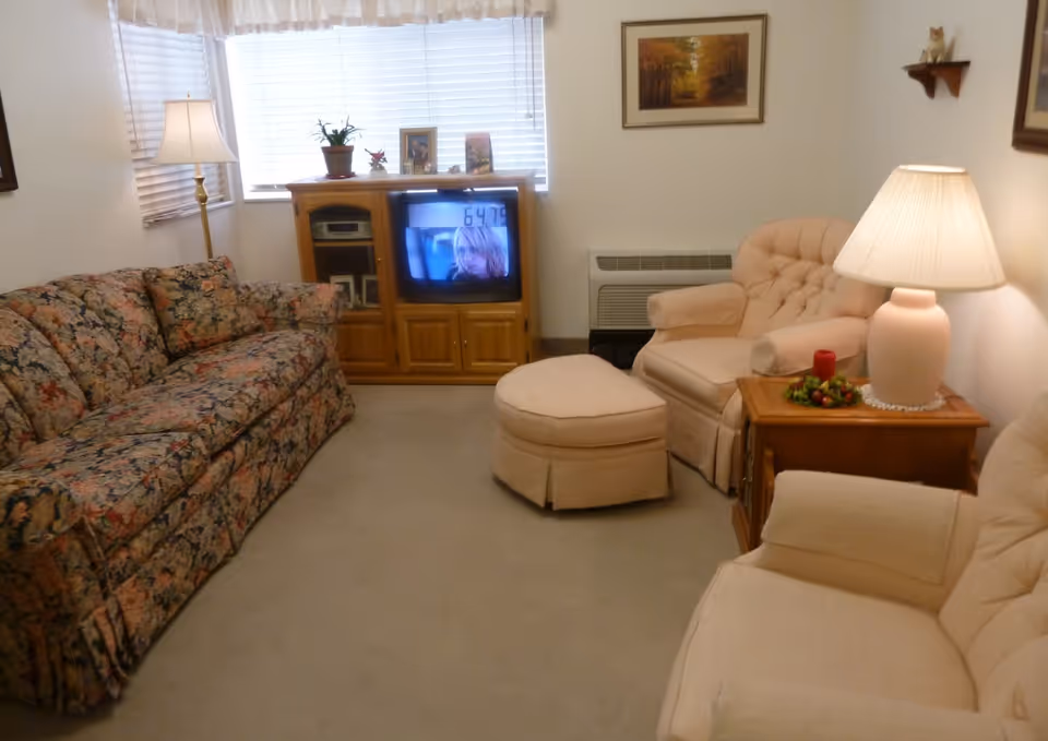 Cozy living room with a floral sofa, two beige armchairs and an ottoman, a wooden TV cabinet, and table lamps.