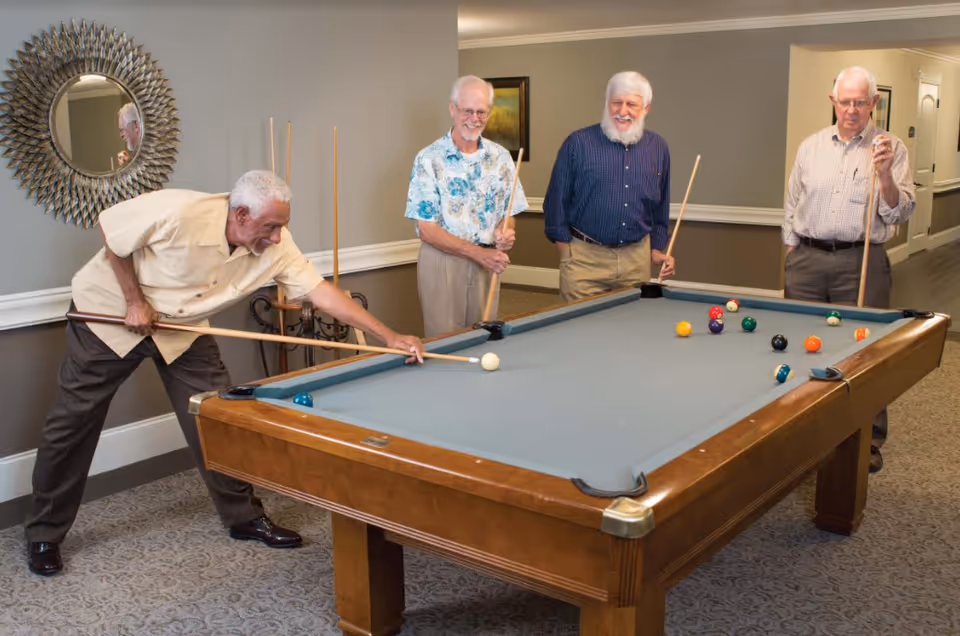 Four elderly men playing pool in a room with beige walls and carpeted floor. One man is taking a shot while the other three watch and hold pool cues. A decorative round mirror is on the wall behind them.