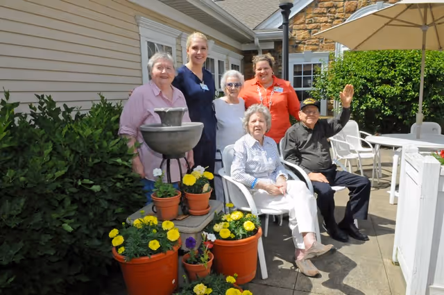 A group of five people, including two elderly women sitting in white chairs and three standing adults, posing outdoors near a building with beige siding. There are several potted yellow and purple flowers and a small water fountain in front of them. One elderly man is sitting and waving. The setting appears to be a patio area with white tables and chairs and an umbrella.
