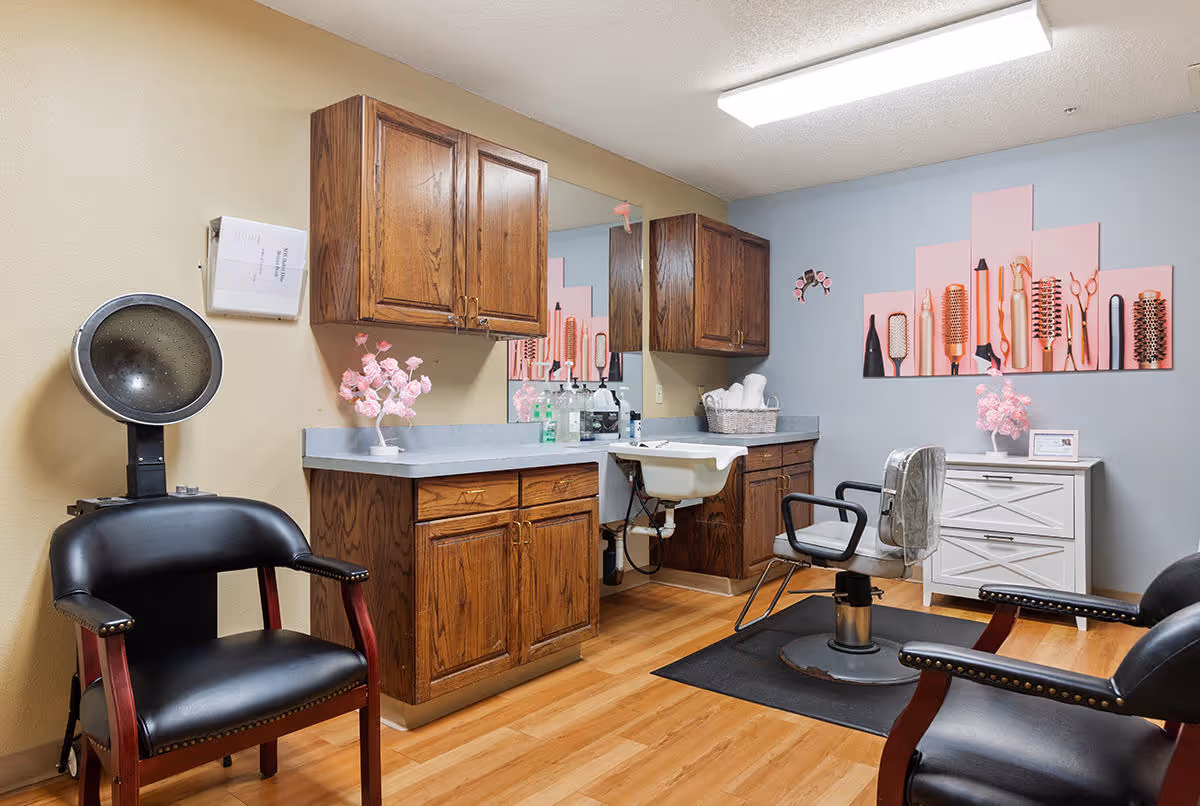 Interior of a hair salon area with wooden cabinets, a sink, a salon chair, a hair dryer chair, and a decorative wall art featuring various hair styling tools. The room has light wood flooring and a combination of beige and blue walls.