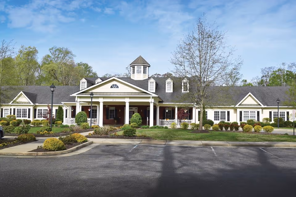 Front exterior view of a single-story senior living facility building with a covered entrance supported by white columns, surrounded by landscaped bushes and trees under a blue sky.