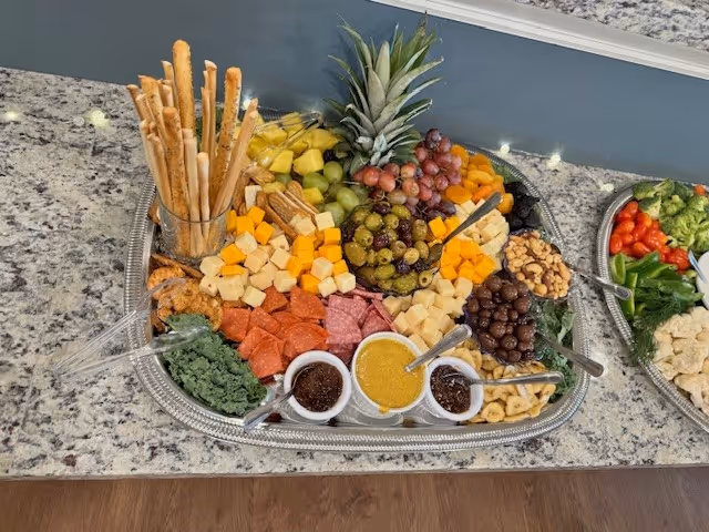 A large silver platter on a granite countertop filled with assorted cheeses, cured meats, olives, fruits, nuts, dips and breadsticks.