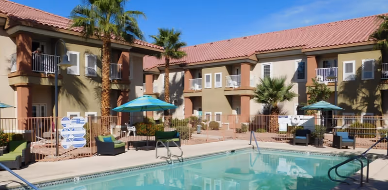 Outdoor swimming pool with lounge chairs, umbrellas, and a two-story residential building with balconies.
