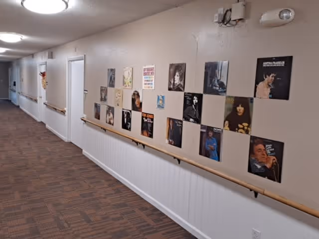A hallway in a senior living facility with brown patterned carpet and beige walls. The walls are decorated with various vintage music album covers arranged in a grid pattern. There are handrails along the wall and several closed white doors along the corridor. Ceiling lights illuminate the hallway.