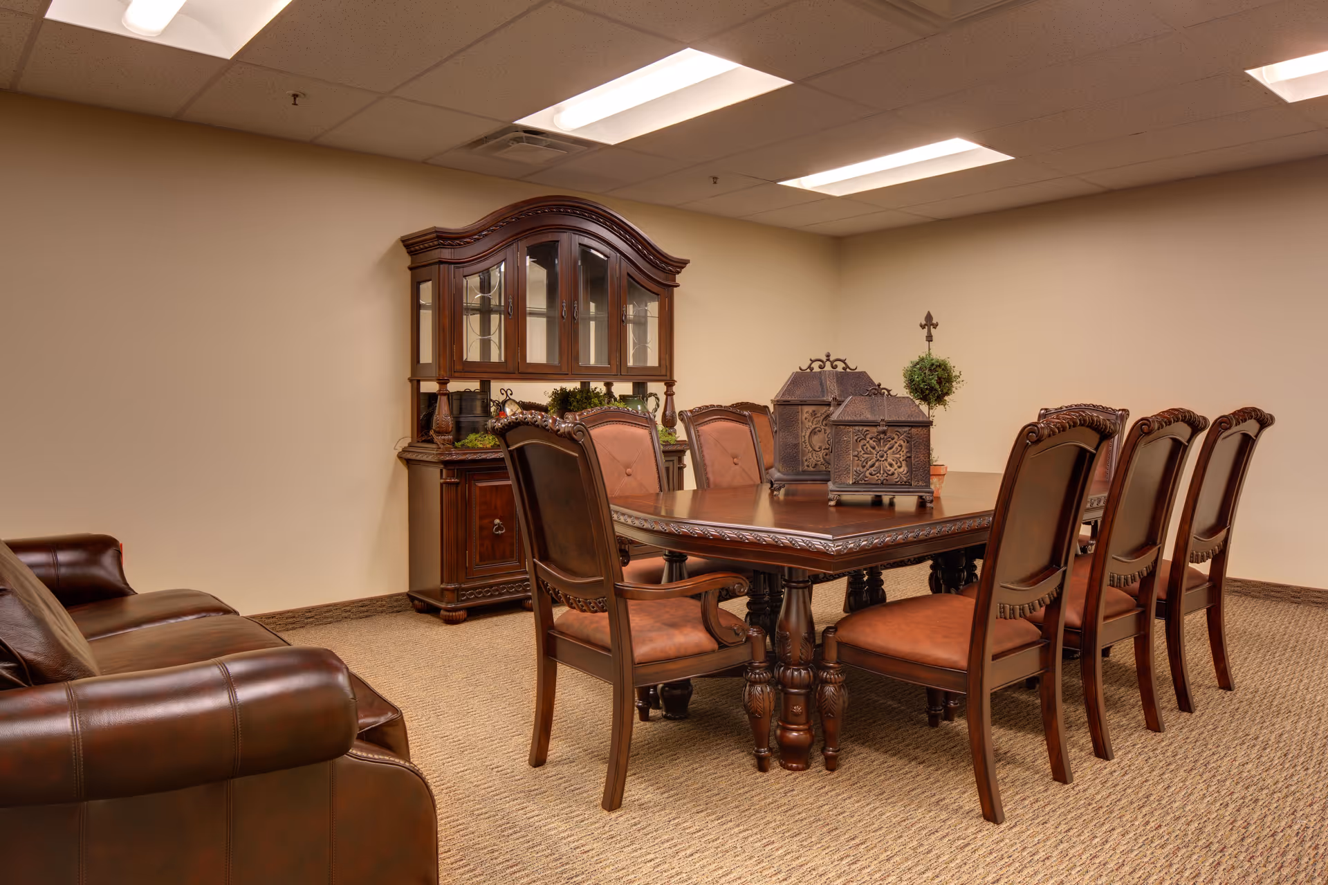 A formal dining room with a large wooden dining table surrounded by eight matching chairs with upholstered seats and backs. There is a wooden china cabinet with glass doors against the wall, decorated with small plants and decorative items. A brown leather sofa is partially visible on the left side of the image. The room has beige walls, a carpeted floor, and fluorescent ceiling lights.