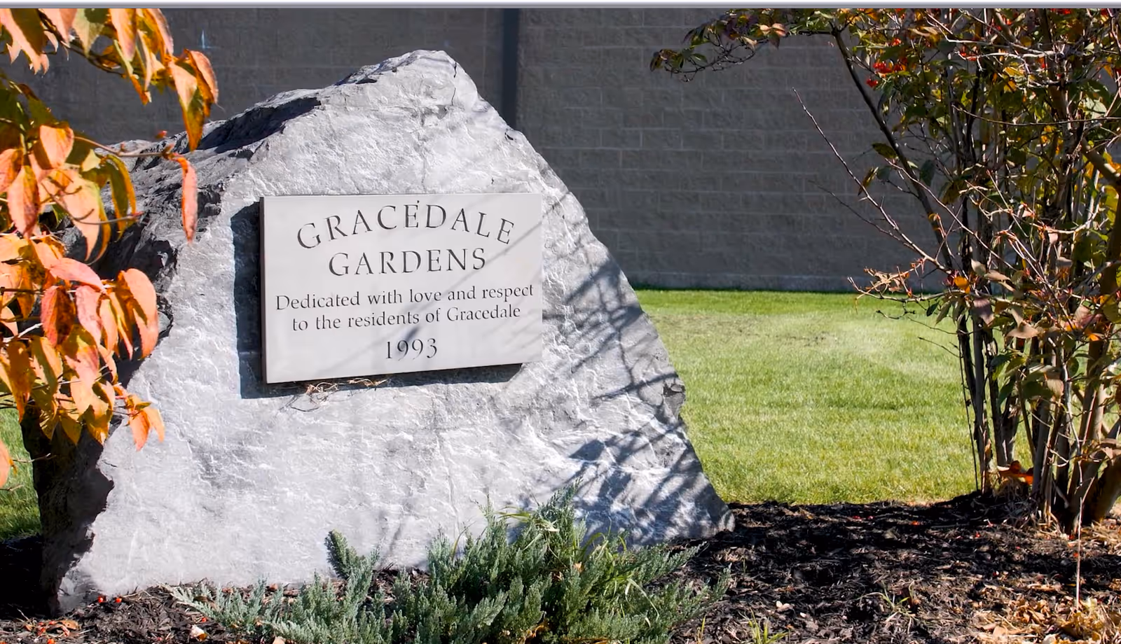 A large gray stone with a plaque that reads 'Gracedale Gardens Dedicated with love and respect to the residents of Gracedale 1993' set in a landscaped garden area with green grass, bushes, and trees with autumn-colored leaves.