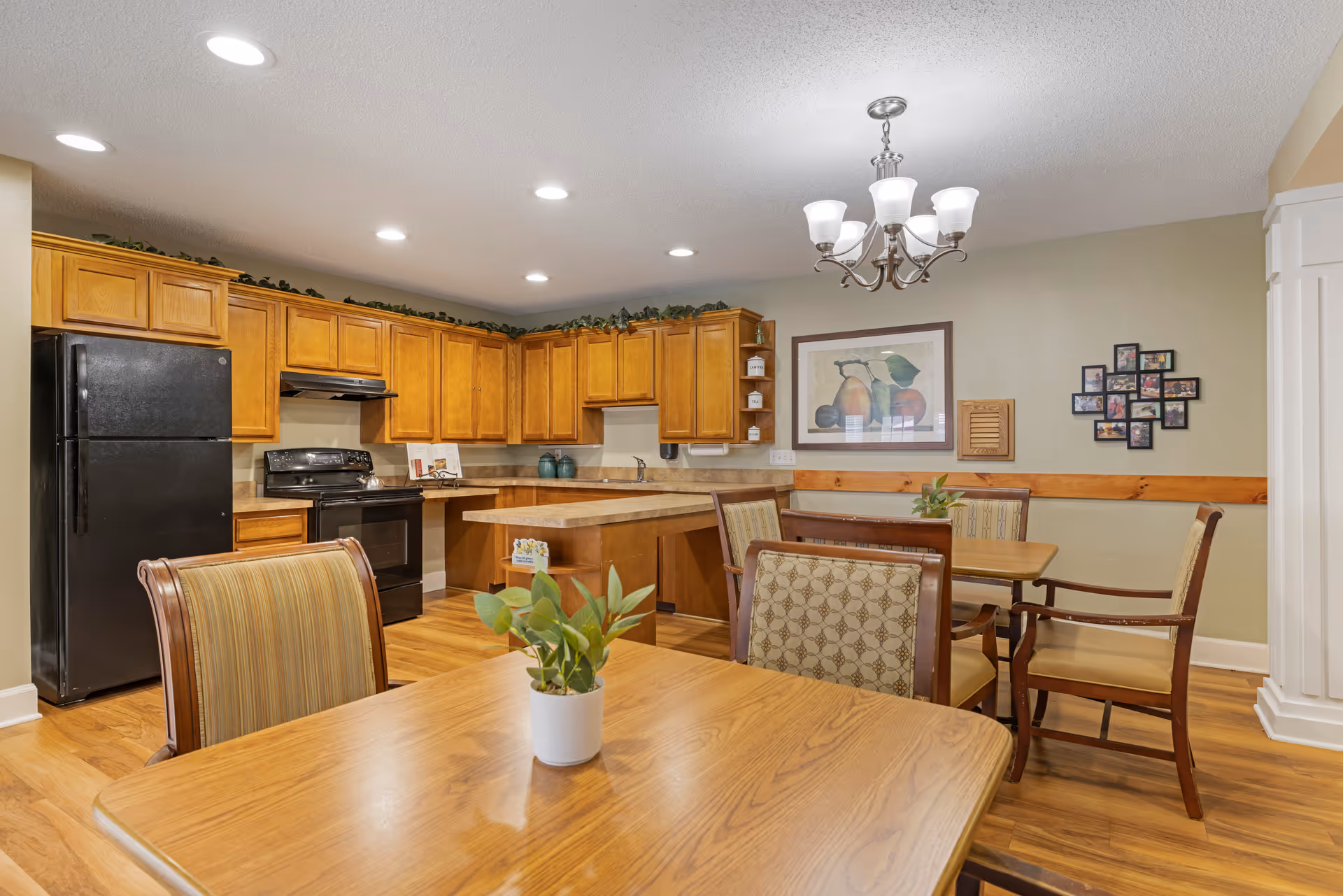 A cozy kitchen and dining area with wooden cabinets, a black refrigerator and stove, and a wooden dining table with chairs. The room has hardwood floors, recessed lighting, a chandelier, and wall decorations including a framed fruit print and a photo collage.