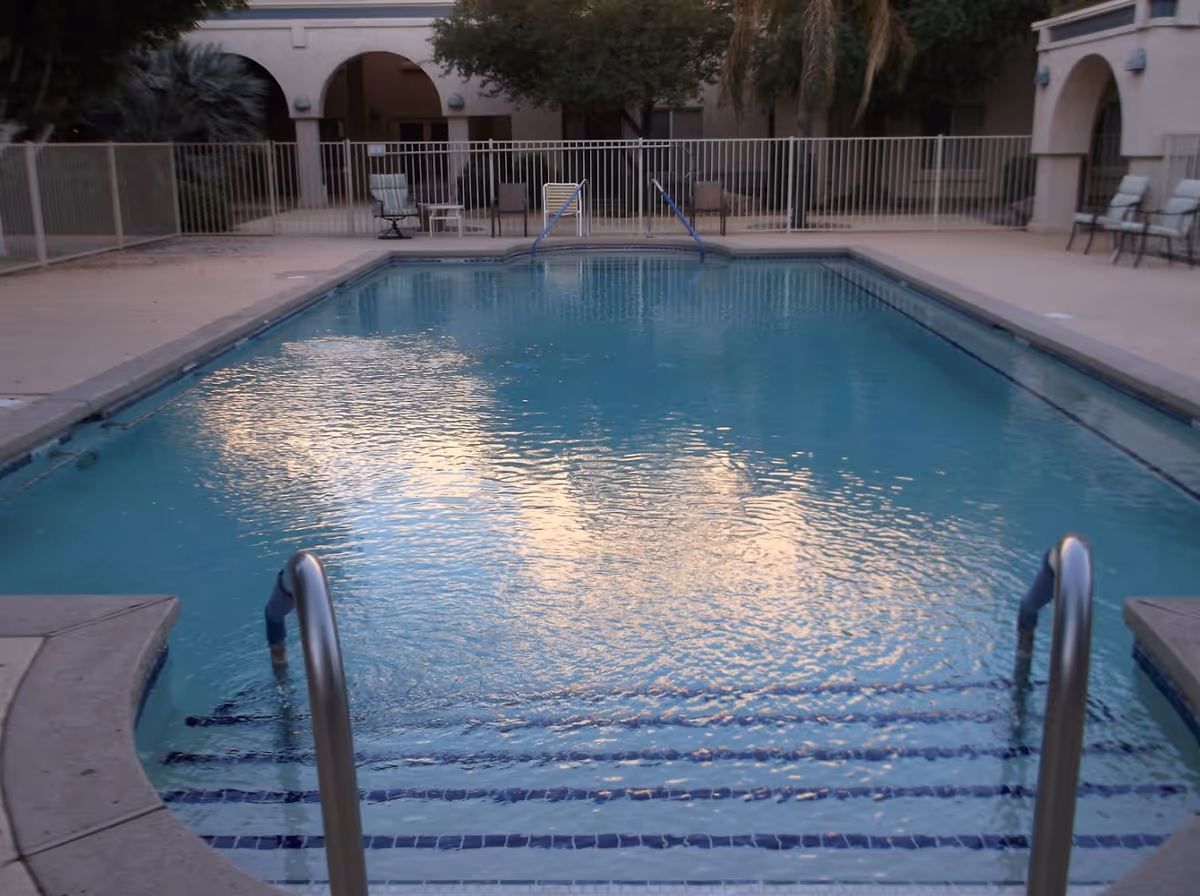 Rectangular outdoor swimming pool in a courtyard with steps and metal handrails, surrounded by a fence and seating near arched building facades.