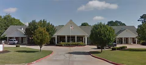 Front exterior view of Reunion Inn Assisted Living facility with a driveway, manicured lawn, trees, and an American flag in front of the building under a partly cloudy sky.