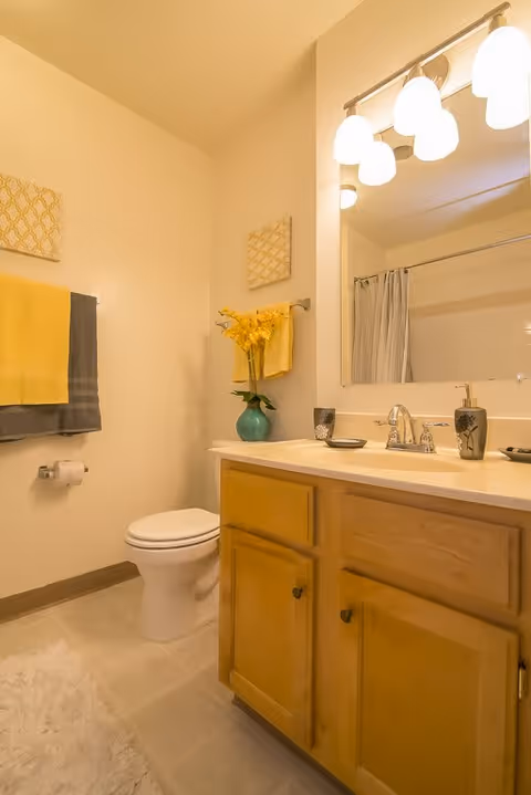 A clean bathroom with a wooden vanity cabinet, a white sink with silver faucet, a large mirror with four light fixtures above it, a toilet, a towel rack with yellow and gray towels, a vase with yellow flowers on the counter, and a shower curtain in the background.