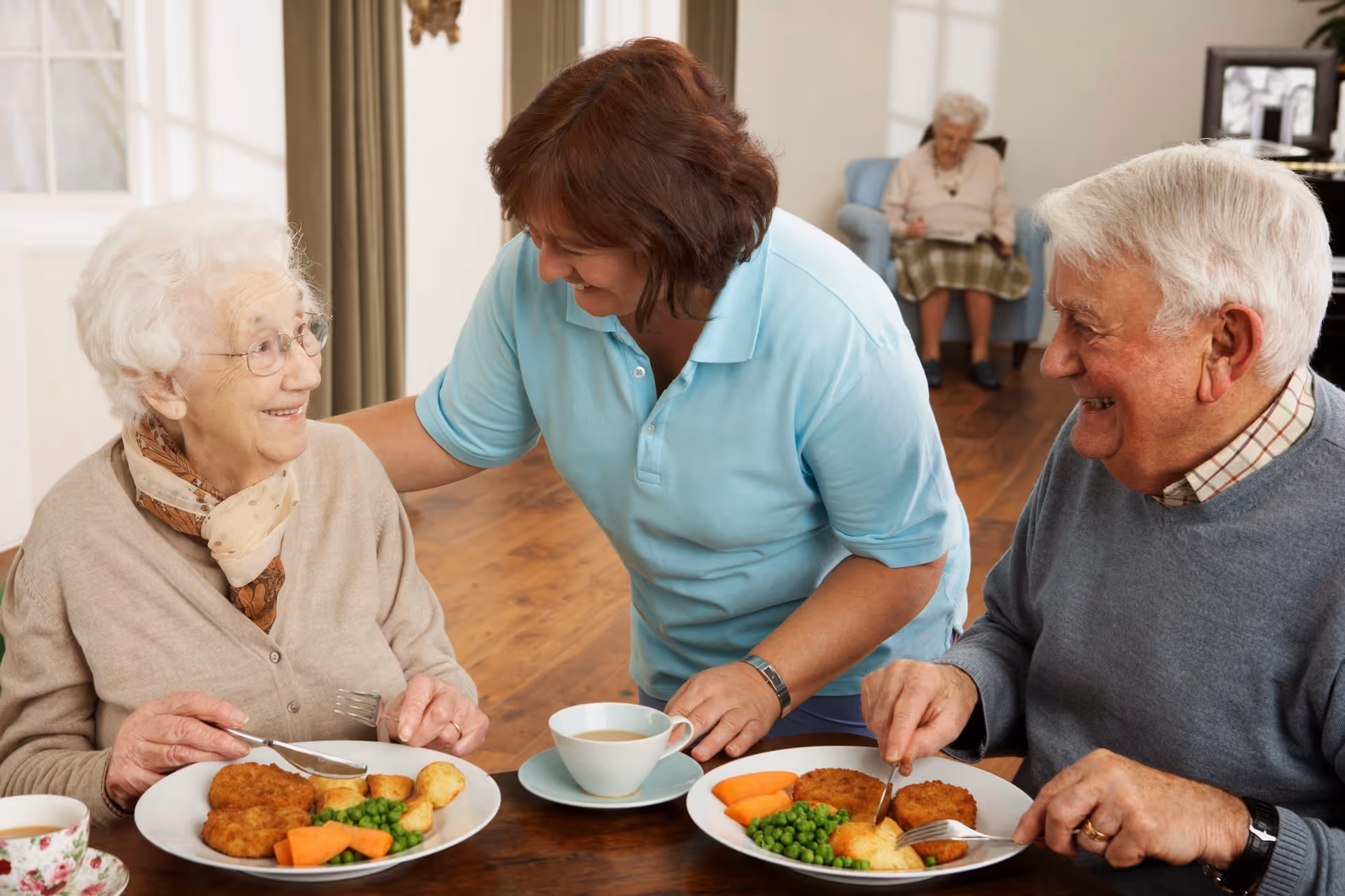 Two elderly people sitting at a dining table with plates of food including peas, carrots, potatoes, and patties, smiling and interacting with a caregiver in a light blue shirt. In the background, another elderly person is seated on a chair reading a book.