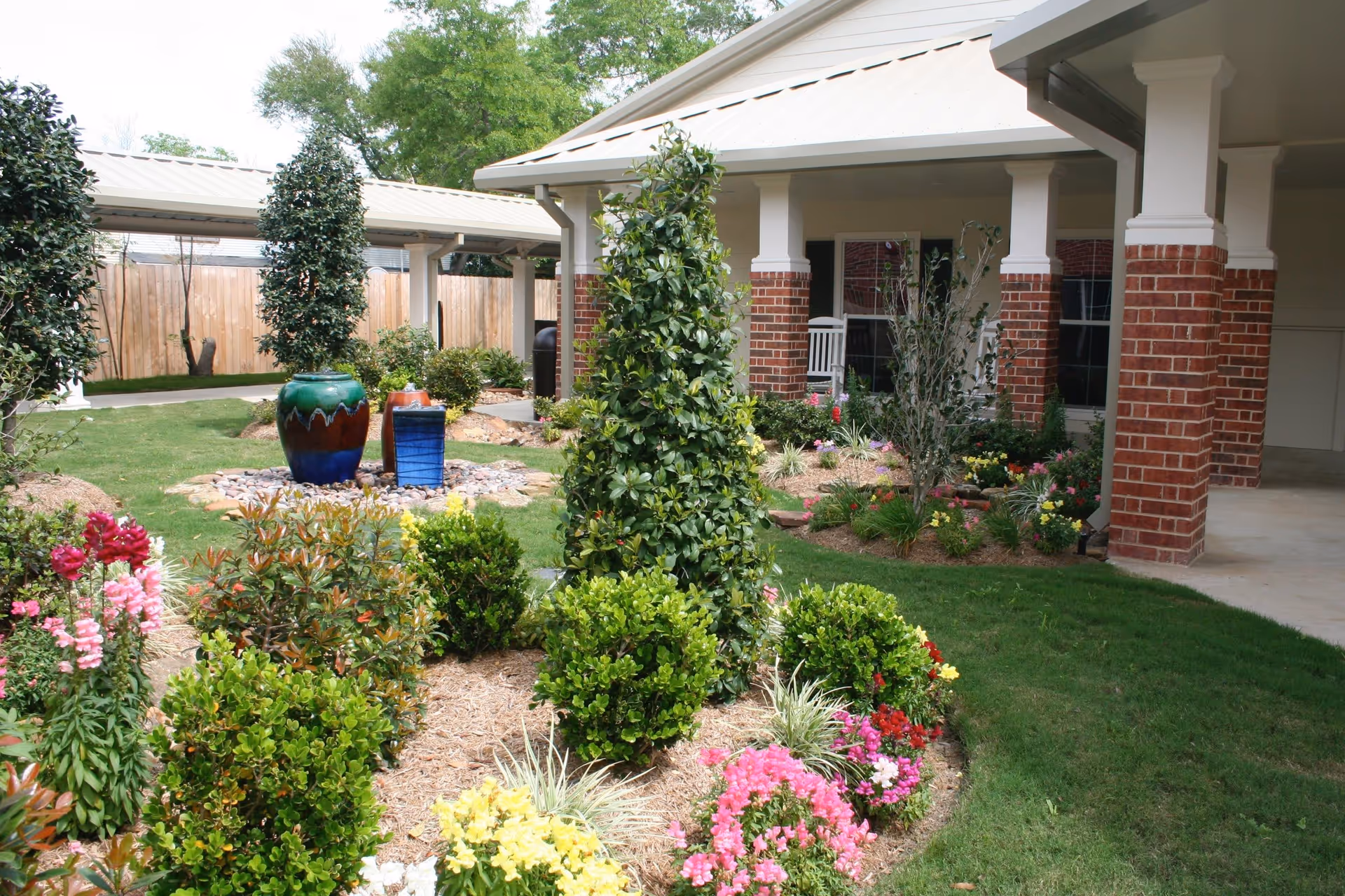A landscaped garden area with various green shrubs and colorful flowers in front of a building with brick pillars and a covered porch. There are decorative ceramic pots and a pathway visible, along with a wooden fence and trees in the background.
