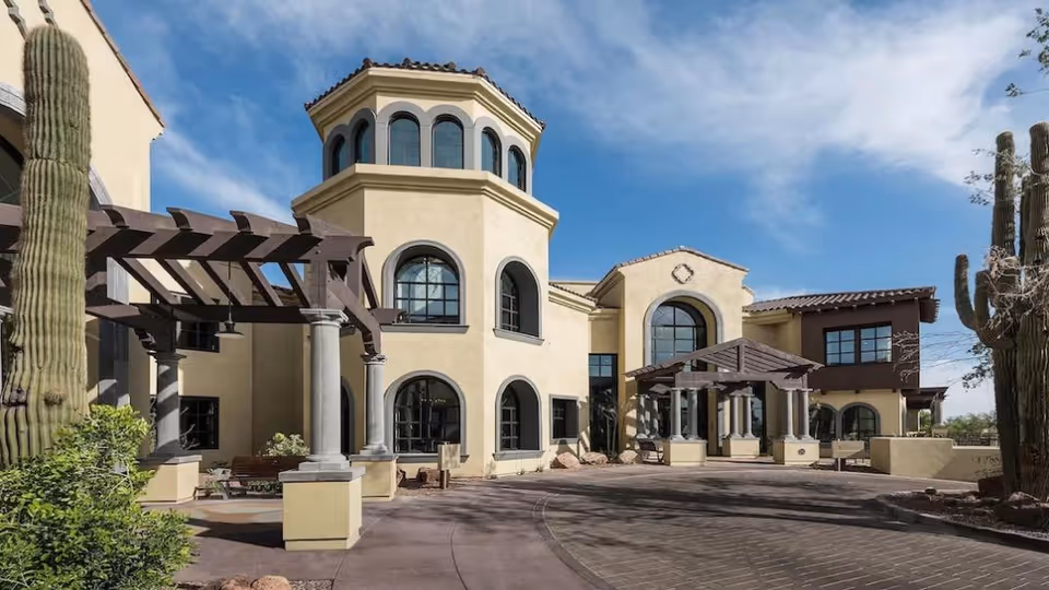 Mediterranean-style senior living facility entrance with a tower, arched windows, pergolas, and desert landscaping including tall cacti.