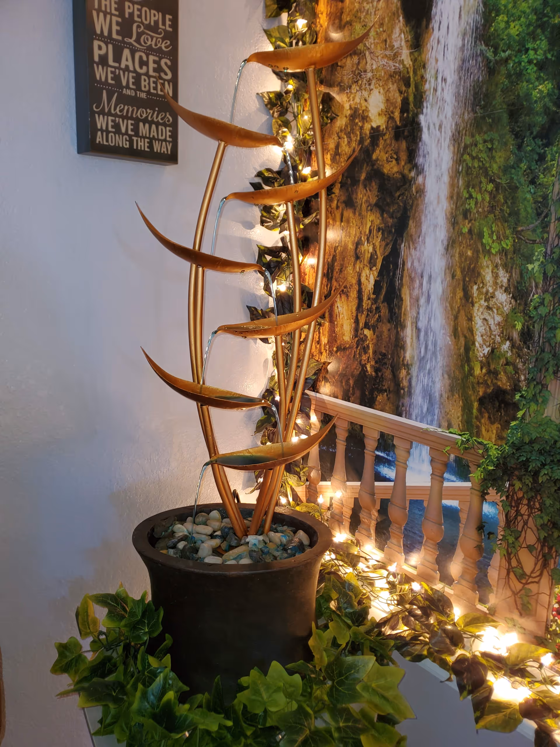 Indoor decorative water fountain with multiple curved spouts pouring water into a pot filled with stones, surrounded by green leafy plants and string lights. A wall mural of a waterfall and a wooden railing is in the background, along with a framed sign that reads 'The people we love, the places we've been, and the memories we've made along the way.'