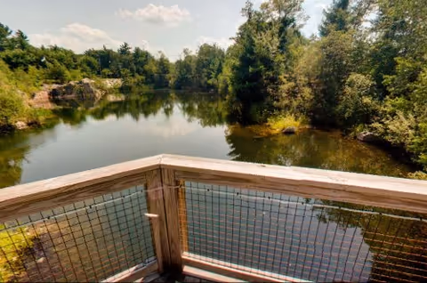 View from a wooden deck railing overlooking a calm pond surrounded by trees and rocky shoreline.