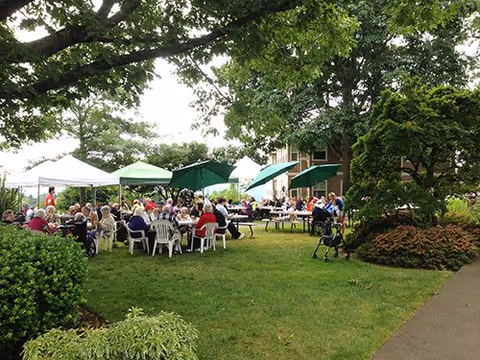 A group of elderly people sitting at tables under white and green canopies in a grassy outdoor area with trees and bushes, enjoying a social gathering at Elite Care at Oatfield Estates.