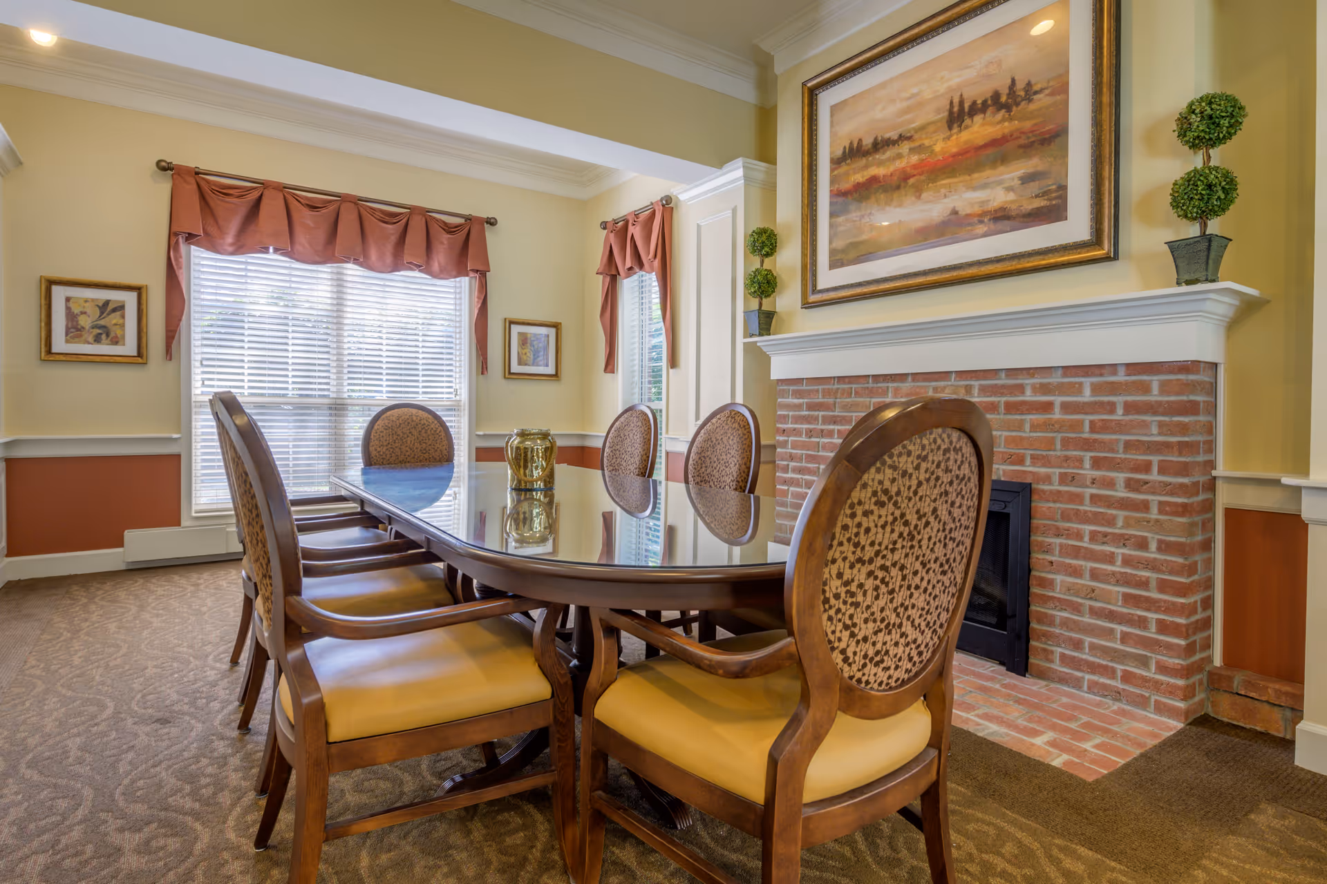 A dining room with a long wooden table surrounded by six upholstered chairs. The room features a brick fireplace with a large framed painting above it and two small potted topiary plants on the mantel. There are two windows with blinds and red valance curtains, along with framed artwork on the walls. The carpet has a patterned design and the walls are painted in warm tones.
