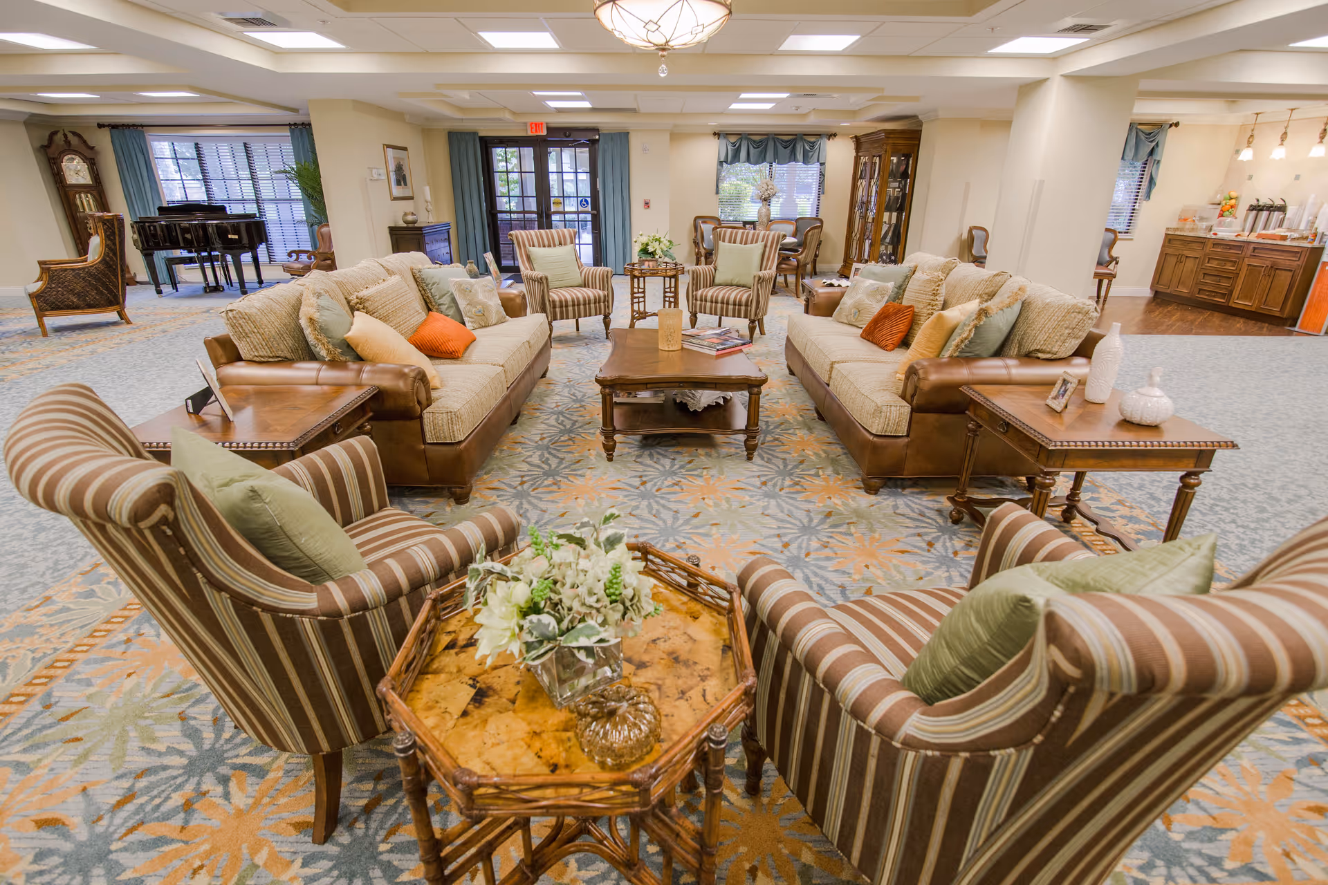 A spacious and well-lit senior living facility common area with two beige sofas adorned with multiple cushions, four striped armchairs, and wooden coffee and side tables. The room features a patterned carpet, a grand piano near the window, a grandfather clock, and a dining area with a wooden cabinet and chairs in the background. Large windows with curtains allow natural light to fill the space.