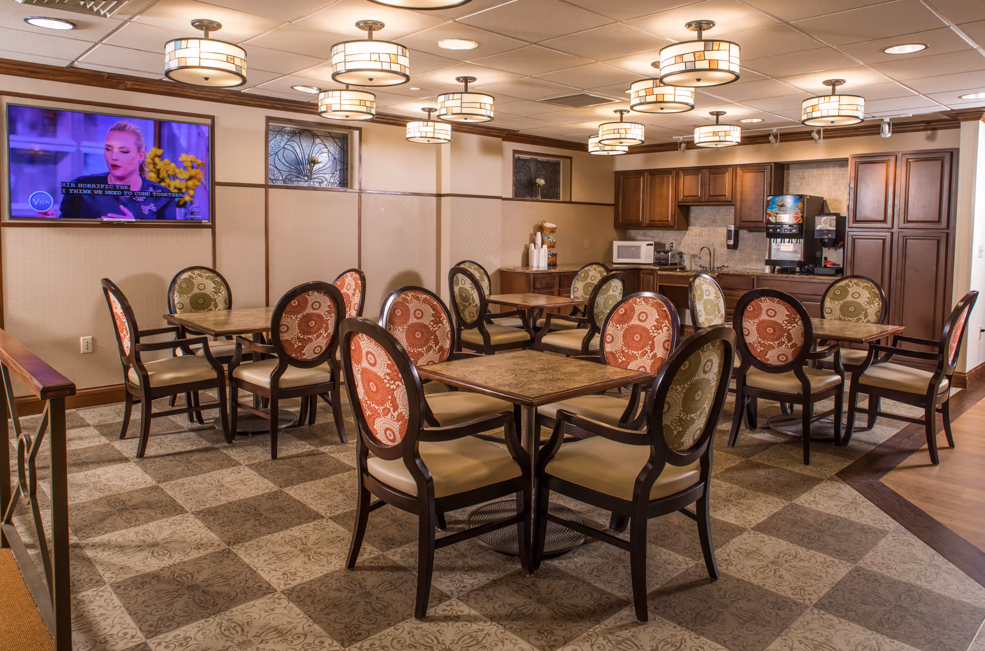 A dining area in a retirement community featuring several square tables with chairs that have patterned backs in red and green. The room has a tiled floor with a decorative pattern, multiple ceiling lights, and a wall-mounted TV showing a woman speaking. There is a kitchenette area with wooden cabinets, a microwave, a coffee machine, and a sink in the background.