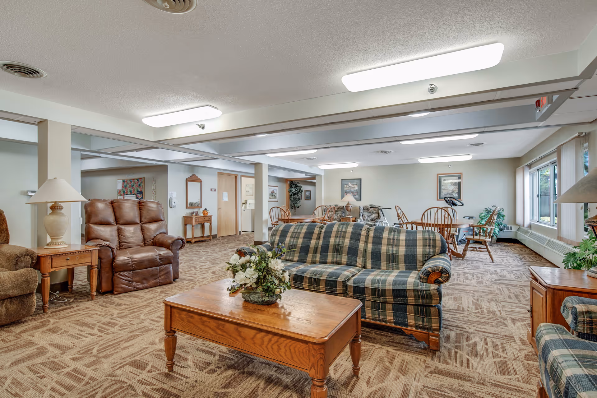 A spacious assisted living common area with patterned carpet, a plaid sofa, a wooden coffee table with a floral arrangement, a brown leather recliner, and several wooden chairs and tables. The room is well-lit with ceiling lights and has large windows with vertical blinds allowing natural light to enter. There are framed pictures on the walls and a few plants adding greenery to the space.