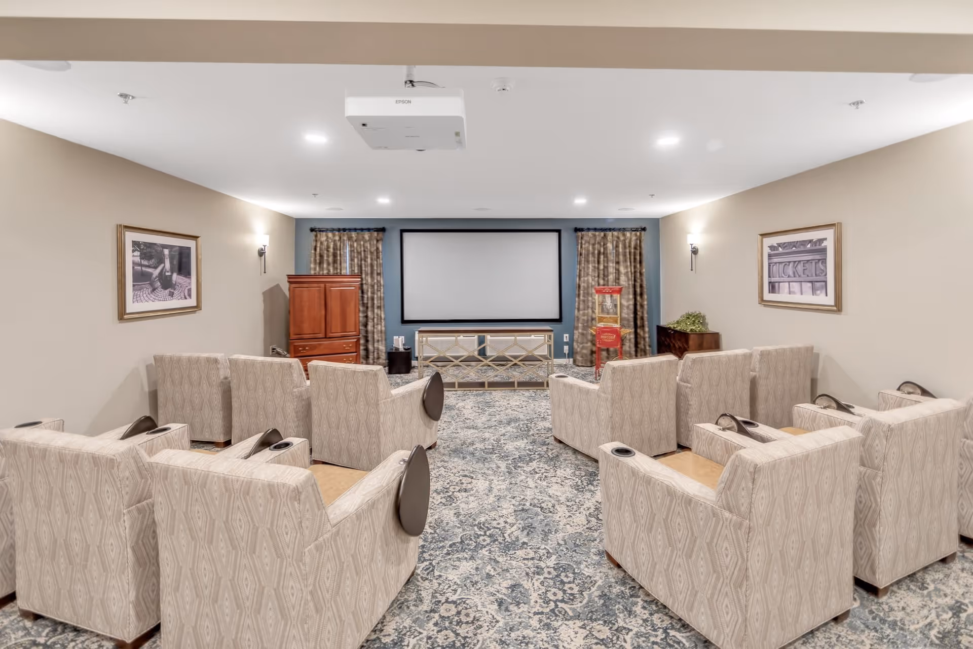 A cozy home theater room with two rows of beige patterned armchairs facing a large projector screen. The room has a patterned carpet, framed pictures on the walls, curtains on the windows, and a popcorn machine near the screen. A ceiling-mounted Epson projector is visible.