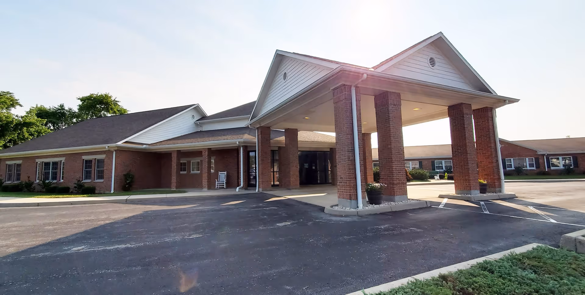 Exterior view of Grace Brethren Village showing a single-story brick building with a covered entrance supported by four brick columns, a paved driveway, and some greenery around the building under a clear sky.