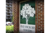 Brick-framed entrance sign with a white tree silhouette on a green background reading "OLD HICKORY TOWERS," with an apartment building and parked cars visible behind it.