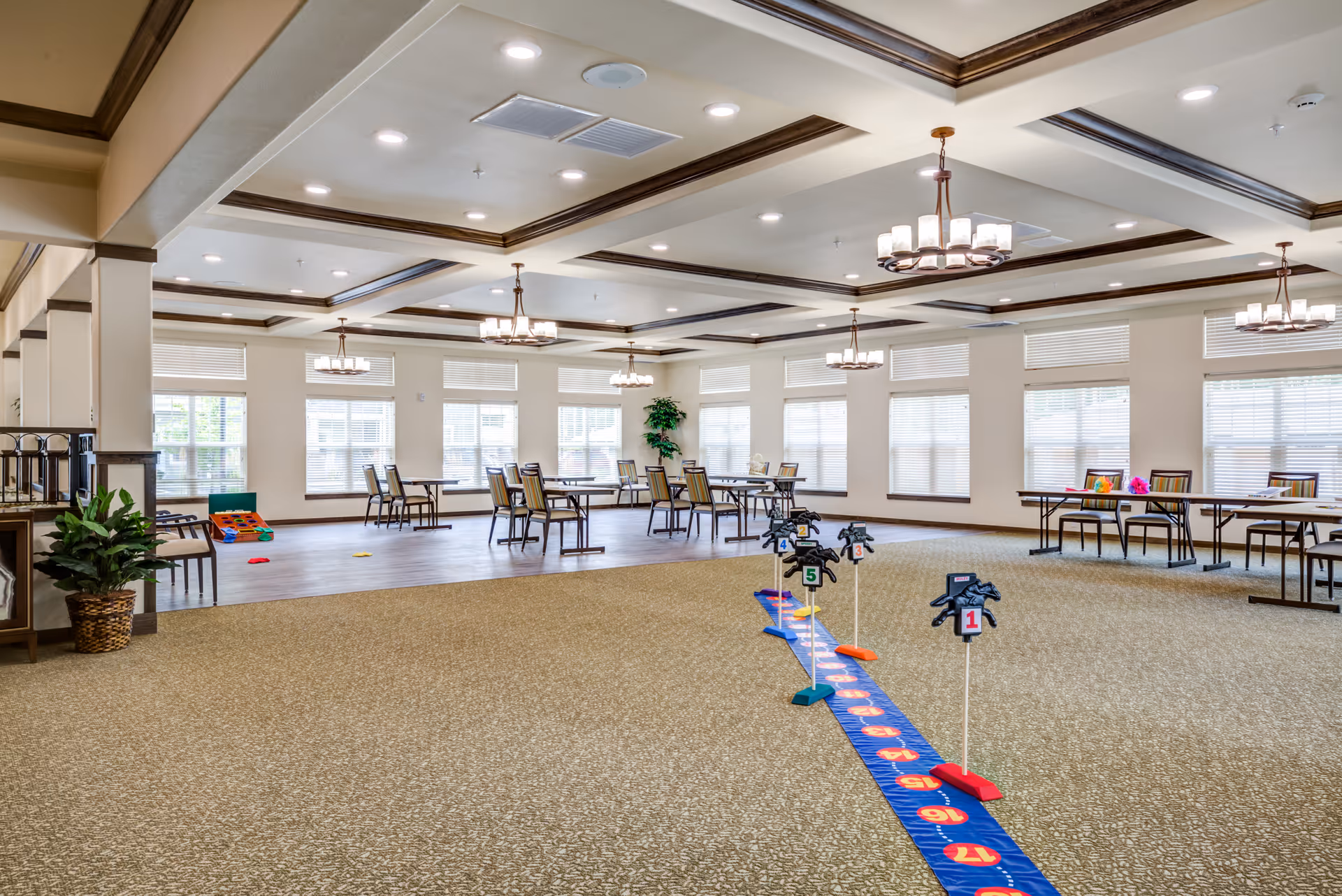 A spacious, well-lit activity room with large windows and multiple tables and chairs arranged around the perimeter. The room features a carpeted area with a colorful game setup involving numbered horse figures on sticks and a blue mat with numbers. The ceiling has recessed lighting and decorative chandeliers, and there are a few potted plants placed around the room.