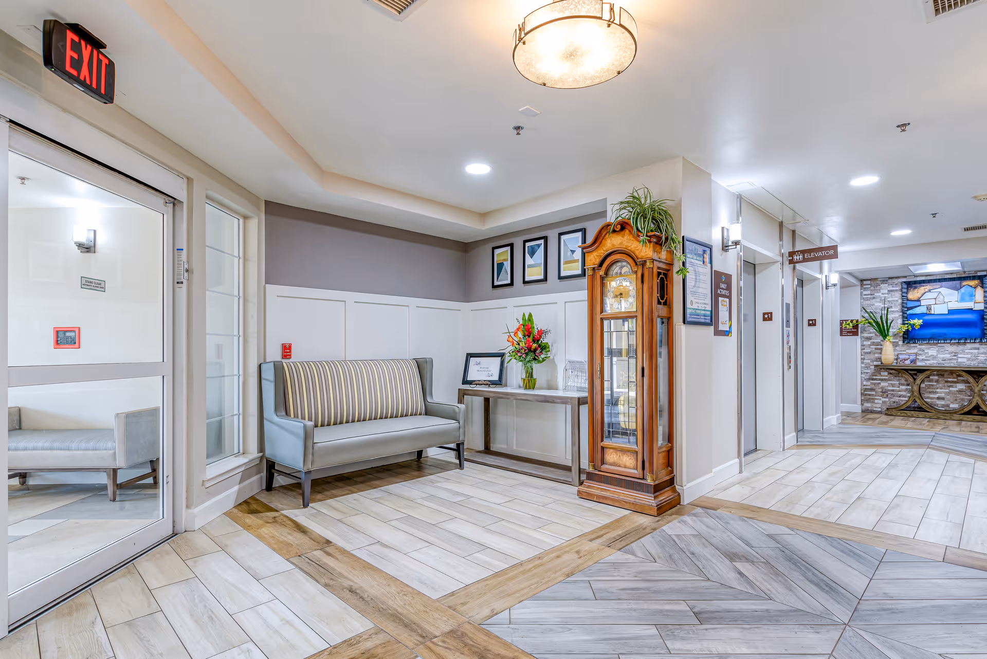 Interior view of a senior living facility lobby area with a striped bench, a wooden grandfather clock, a table with a flower arrangement and framed pictures on the wall. There are two elevators on the right side and a TV mounted on a stone accent wall in the background. The floor has a mix of light wood and tile patterns, and an exit door is visible on the left.