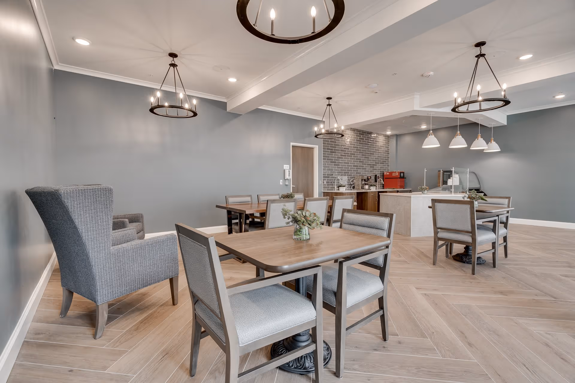 A modern dining area in a senior living facility with wooden tables and cushioned chairs. The room features light wood flooring arranged in a herringbone pattern, gray walls, and multiple hanging light fixtures including chandeliers and pendant lights. There is a small counter area with coffee machines and a red appliance against a gray tiled wall in the background.