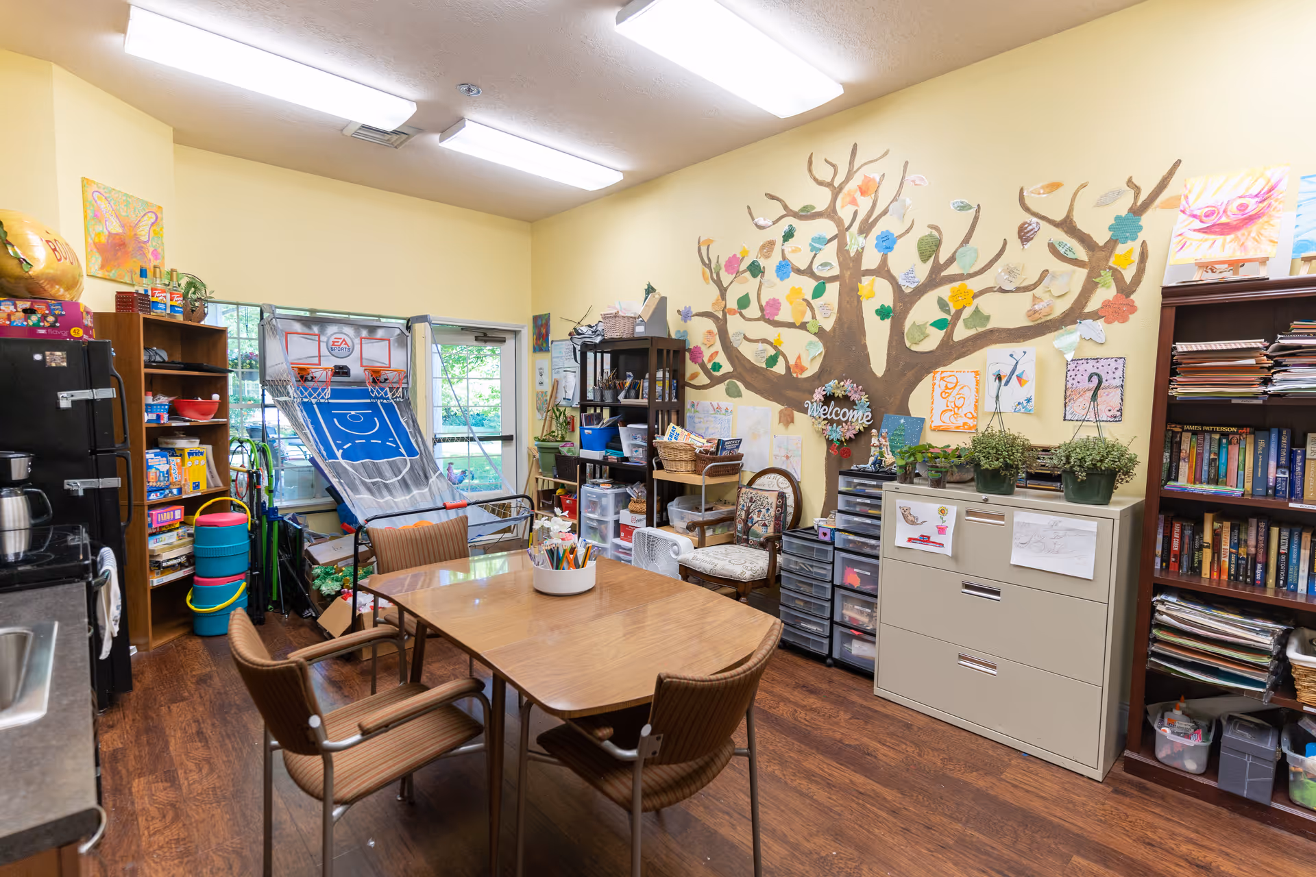 A brightly lit room with a wooden table and four chairs in the center. The walls are painted yellow and decorated with a large tree mural adorned with colorful paper leaves and various artwork. Shelves filled with books, games, and craft supplies line the walls. A basketball arcade game is positioned near the window, and a filing cabinet with plants and drawings on it is also visible.