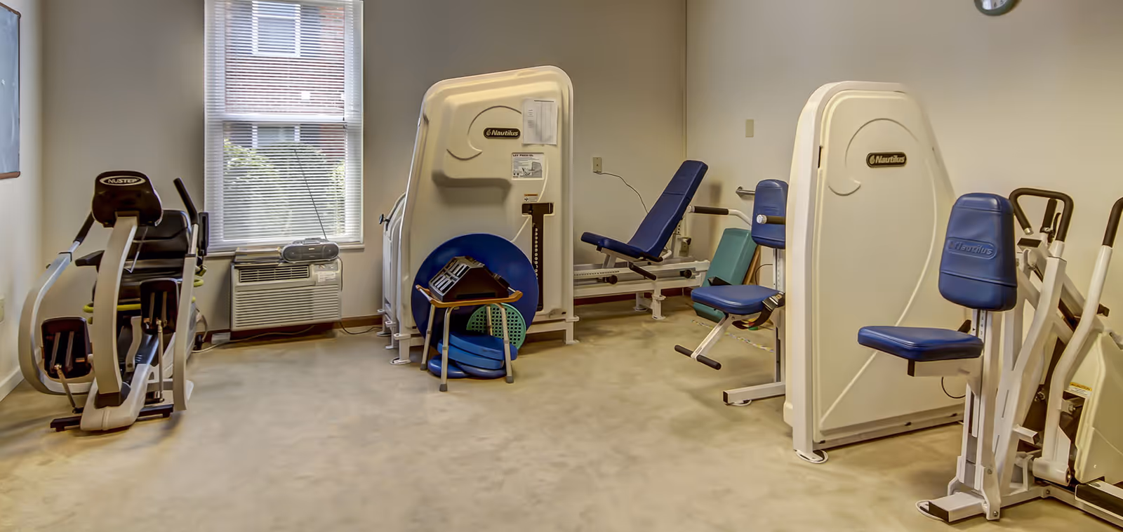 A small fitness room with exercise machines including a recumbent bike, leg press machines, and other strength training equipment. There is a window with blinds, an air conditioning unit below it, and some exercise accessories like balance discs and a small step stool stacked in the corner.