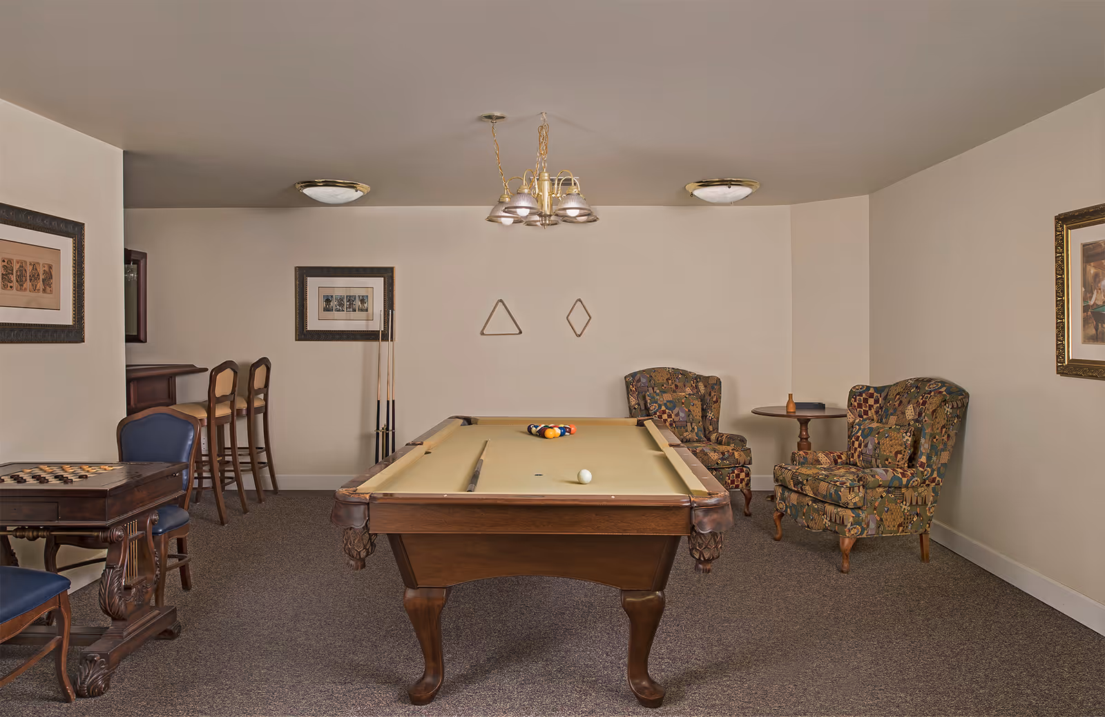 A recreation room with a pool table centered, cue sticks, bar stools, a game table, and upholstered chairs.