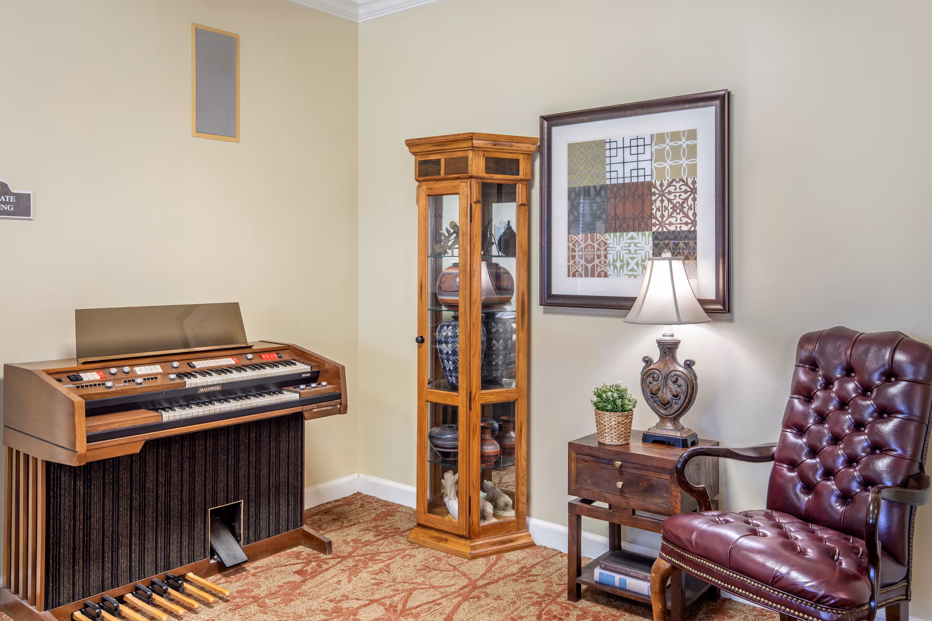 A cozy corner of a room featuring a vintage organ, a wooden glass display cabinet with decorative pottery, a small wooden side table with a lamp and a potted plant, and a burgundy tufted leather armchair. The walls are light beige and there is a framed abstract artwork hanging above the side table.