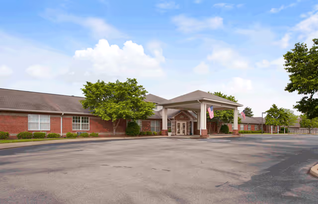 Exterior view of Wellington Manor Jackson, a single-story brick building with a covered entrance supported by white columns. There are American flags hanging near the entrance, trees and shrubs around the building, and a large empty parking lot in front under a partly cloudy sky.