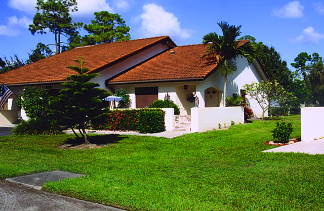 Single-story residential building with a red-tiled roof, white exterior walls, and a small front yard with green grass, bushes, and palm trees under a blue sky with some clouds.