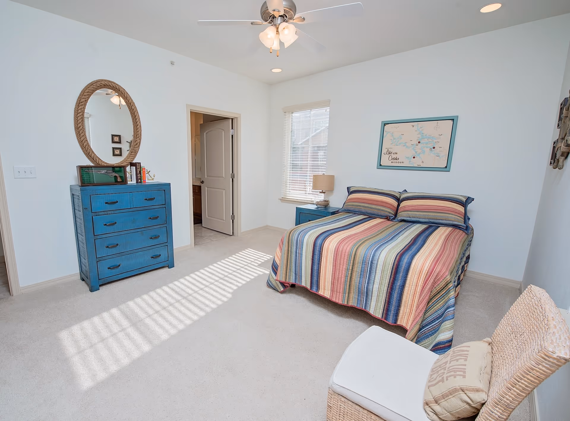 A bright bedroom with a striped multicolored bedspread on a double bed, a blue dresser with a round mirror above it, a window with blinds letting in sunlight, a nightstand with a lamp, and a wicker chair with a cushion.