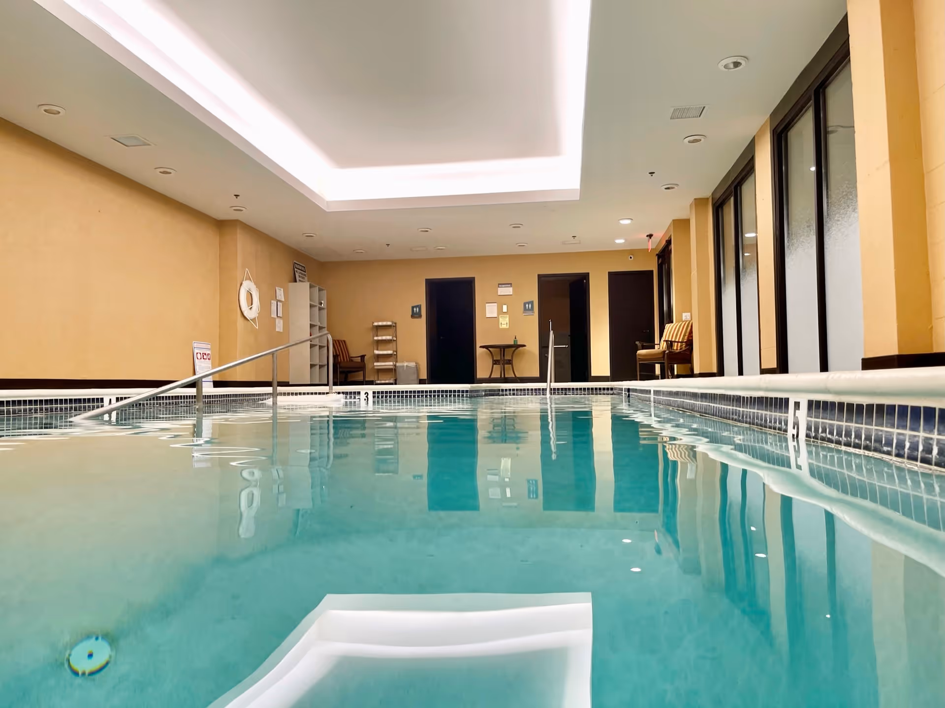 Indoor swimming pool with clear blue water, beige walls, and a white ceiling with recessed lighting. There are chairs and small tables along the walls, and doors at the far end of the room. A metal handrail leads into the pool.