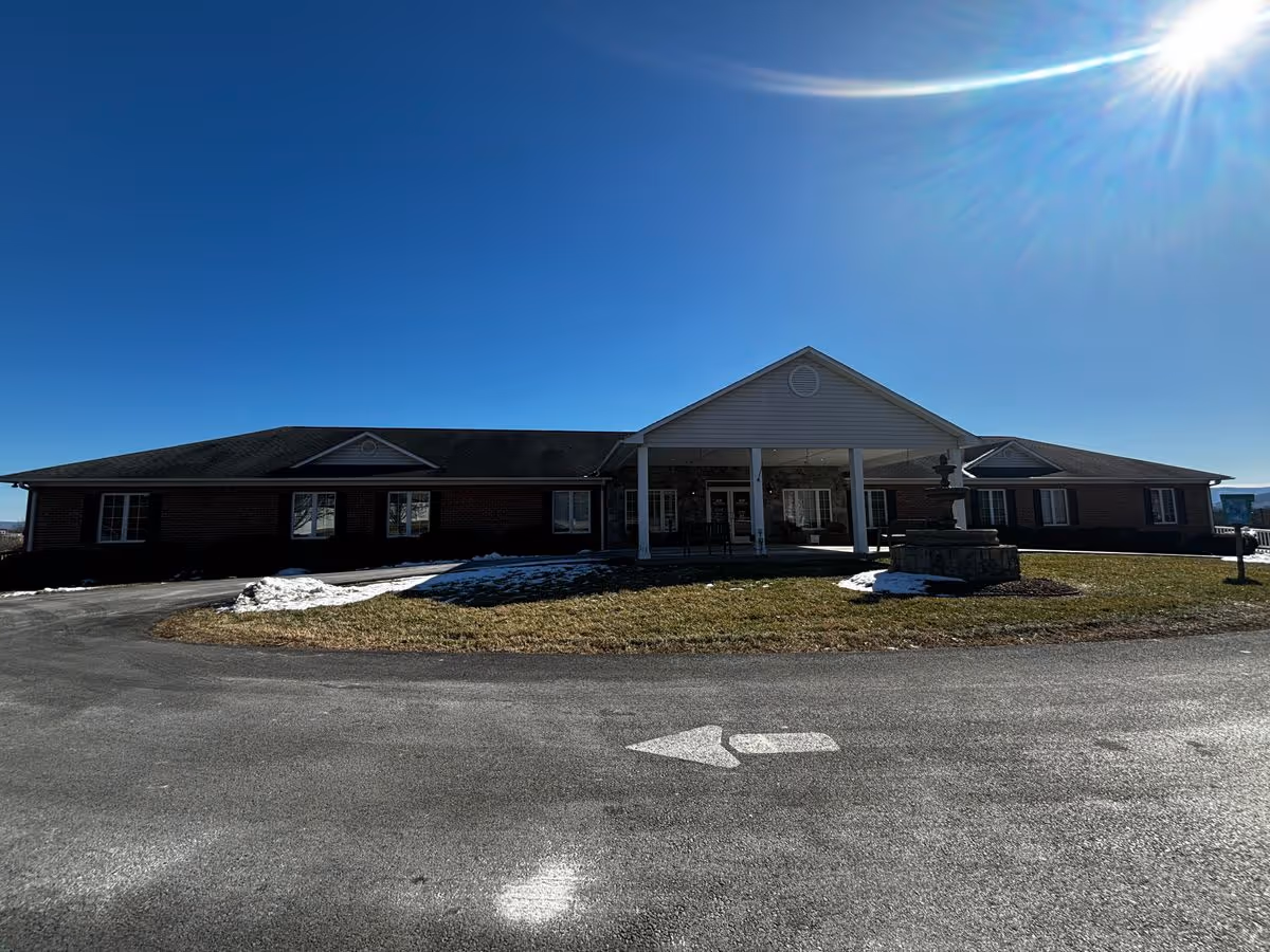 Front view of a single-story brick senior living building with a covered portico, circular driveway, and small fountain under a clear blue sky.