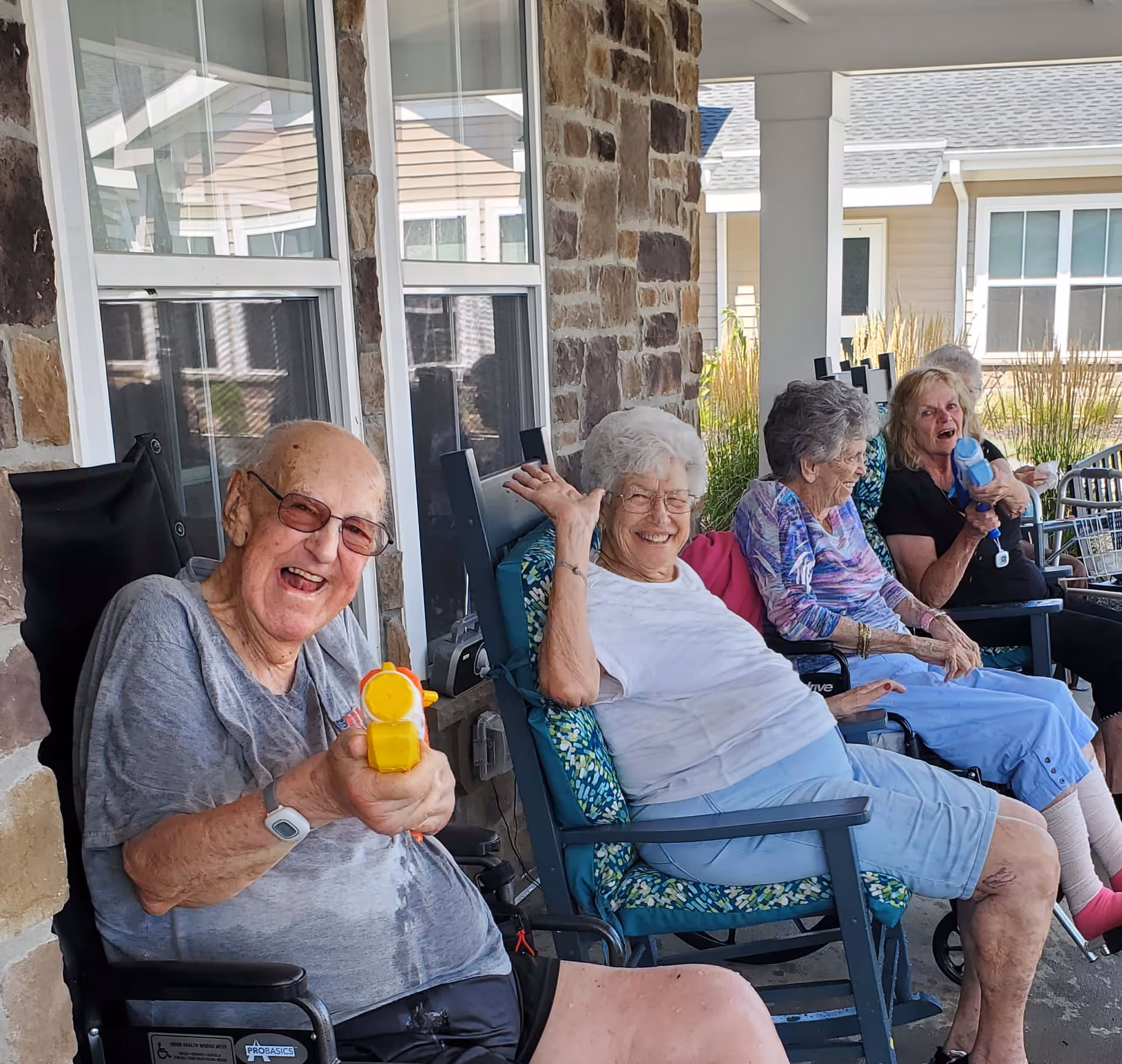 A group of elderly people sitting outside on a porch in chairs and wheelchairs, smiling and enjoying themselves. One man in the foreground is playfully aiming a yellow toy water gun towards the camera. The porch has stone walls and windows, with a residential building visible in the background.