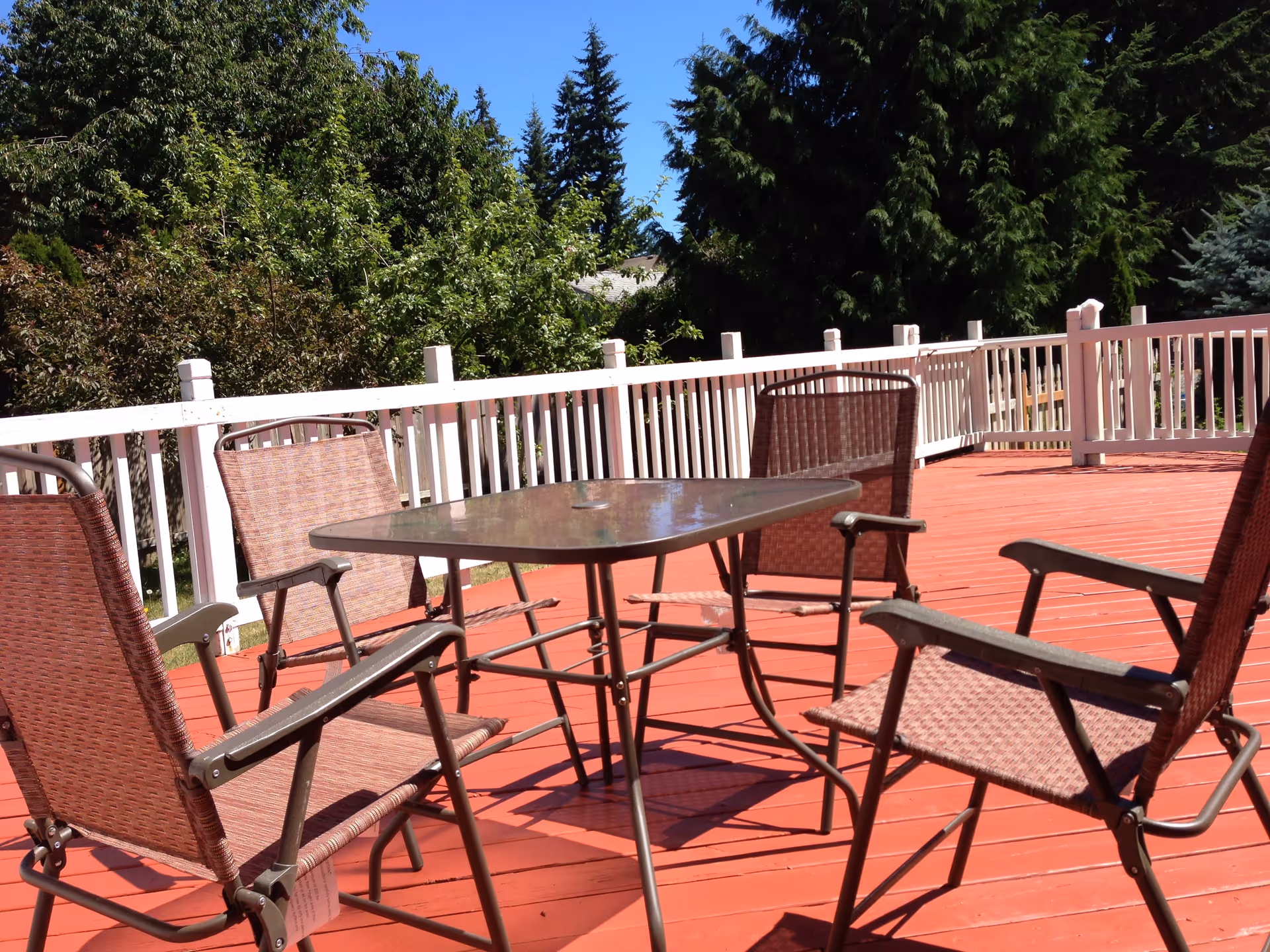 Outdoor deck with a glass-top table and four wicker chairs on a red painted wooden deck, white railing and trees in the background.