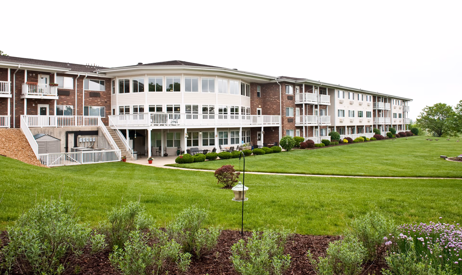 Wide exterior view of a multi-story assisted living facility with brick and white siding, balconies, large windows, and a well-maintained green lawn with shrubs and a bird feeder in the foreground.