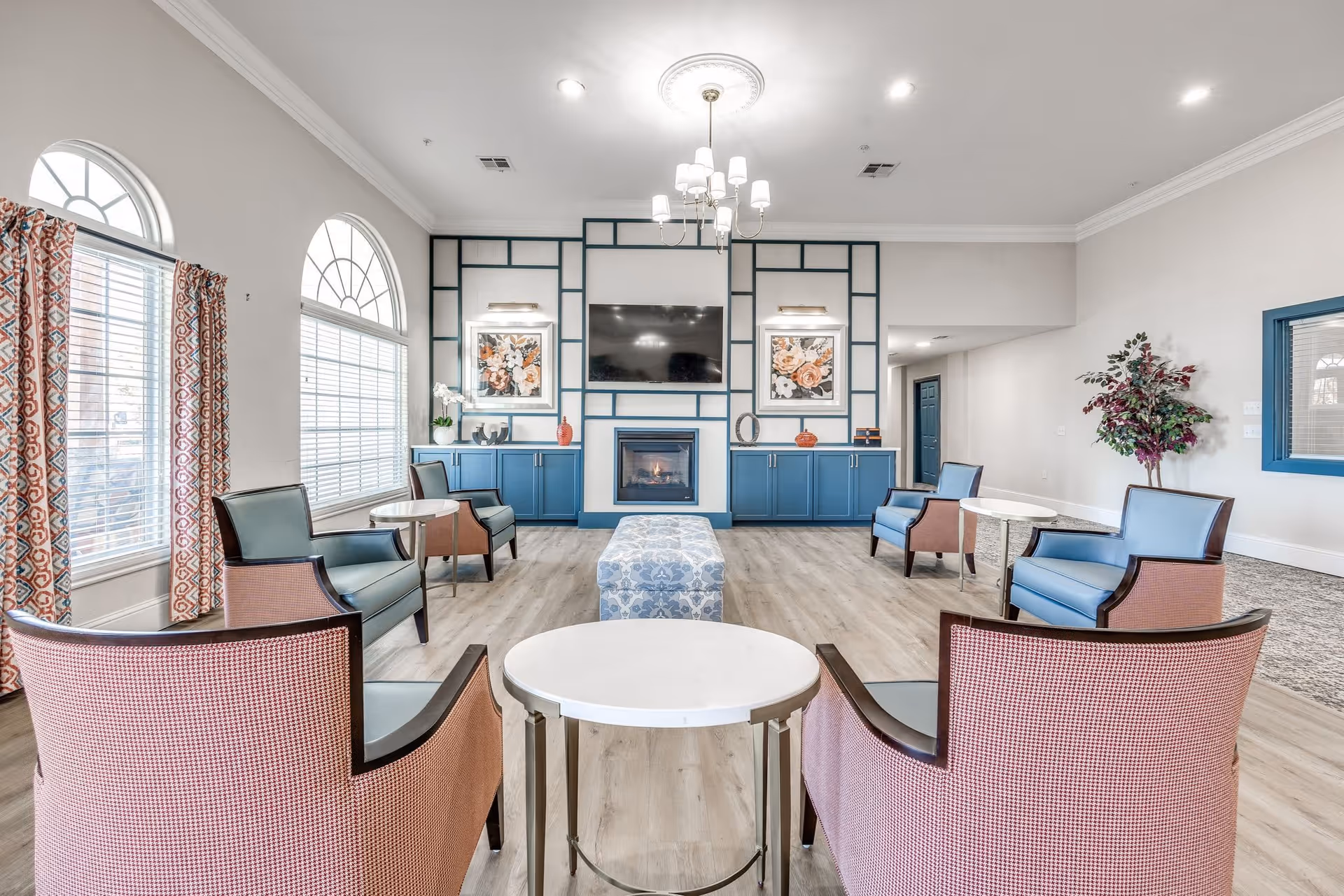 A bright and spacious senior living common area with multiple armchairs arranged around small round tables. The room features large arched windows with patterned curtains, a modern fireplace with a mounted flat-screen TV above it, and blue cabinetry with decorative artwork on the wall. A chandelier hangs from the ceiling, and there is a potted plant in the corner.