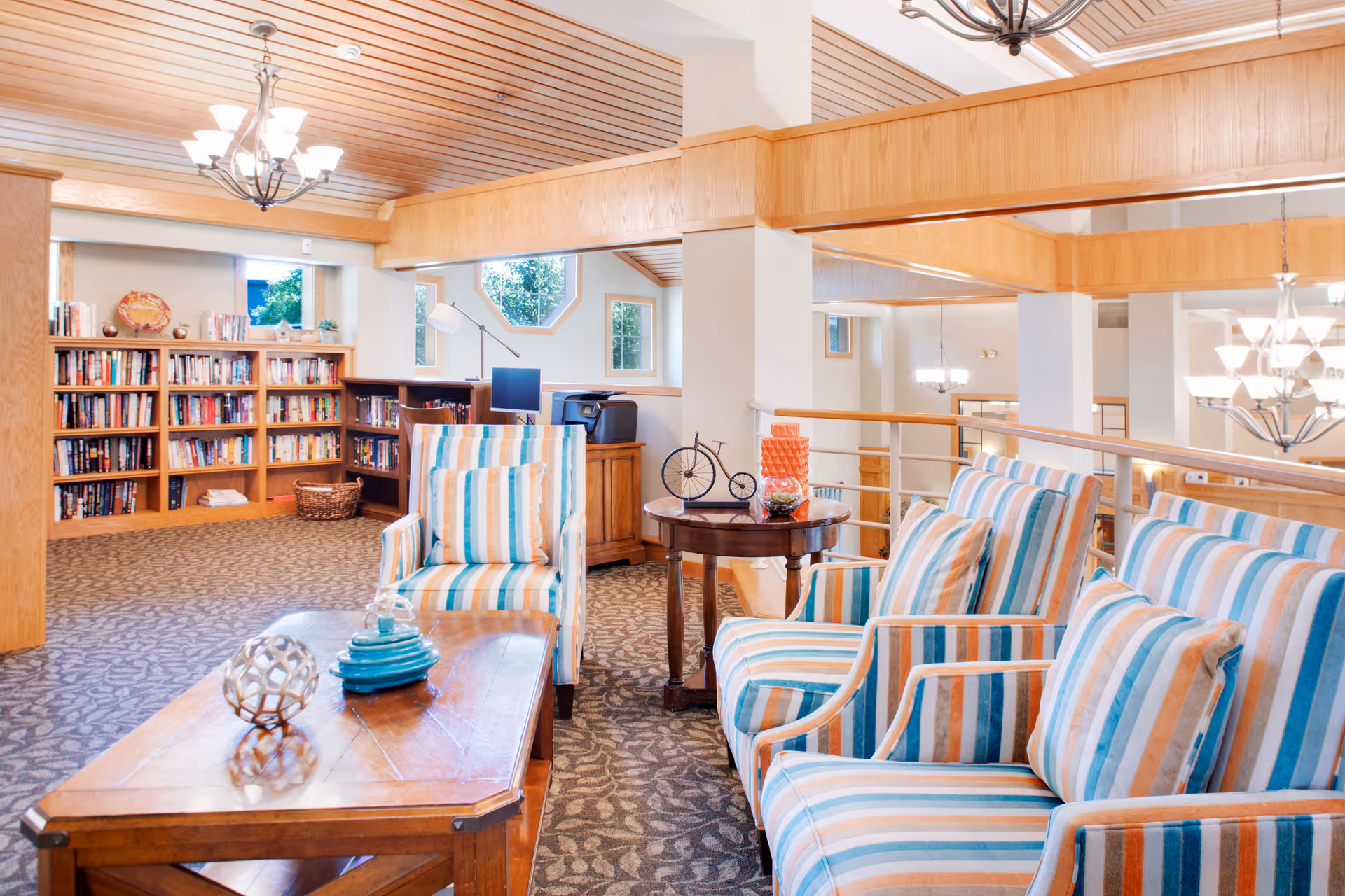 A bright and cozy living room area with striped armchairs in shades of blue, orange, and white arranged around a wooden coffee table. The room features a patterned carpet, wooden ceiling panels, and bookshelves filled with books along the back wall. There are windows letting in natural light and decorative items on the tables and shelves.