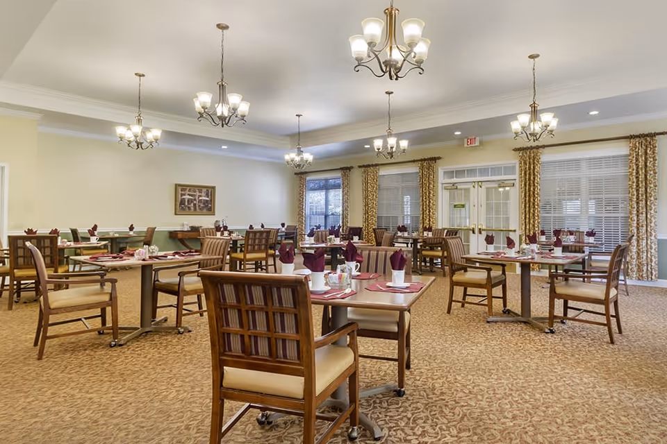 A spacious dining room with multiple wooden tables and chairs arranged neatly. Each table is set with white cups, silverware, and maroon napkins folded upright. The room has beige walls, patterned carpet, large windows with curtains, and several chandeliers hanging from the ceiling providing warm lighting.