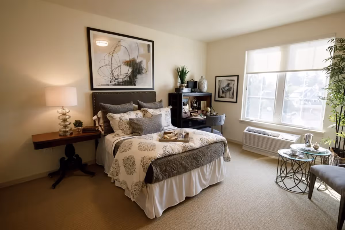 A cozy bedroom with a neatly made bed featuring patterned bedding and multiple pillows. To the left of the bed is a wooden side table with a glass lamp and small plant. Above the bed hangs a large abstract painting. On the right side of the room, there is a window with white blinds, a small round glass table with a tea set, a gray chair, and a tall plant in the corner. A dark wooden desk with a chair and decorative items is positioned against the far wall.