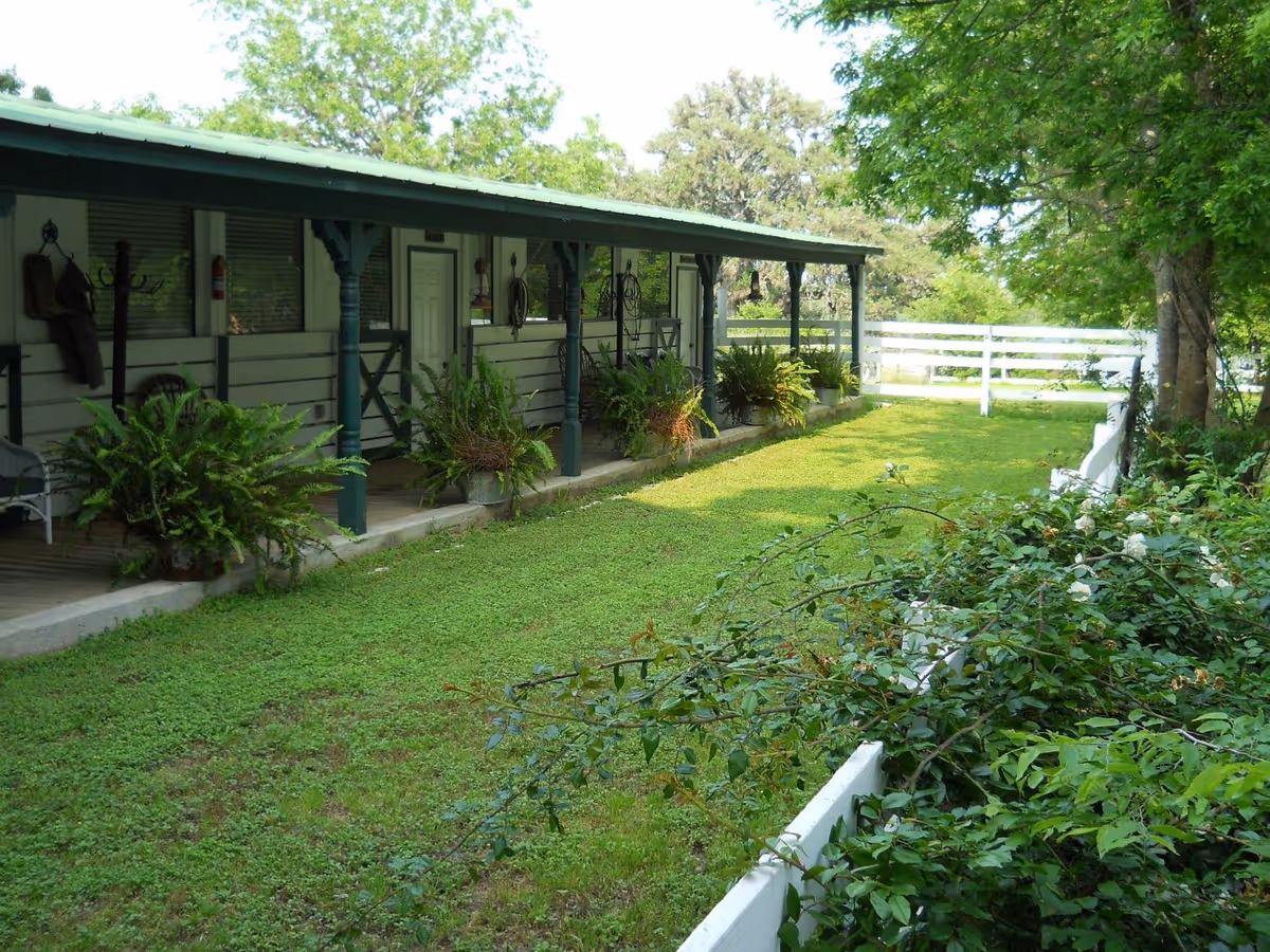 A long single-story building with a covered porch facing a grassy lawn and white fence, surrounded by potted plants and trees.